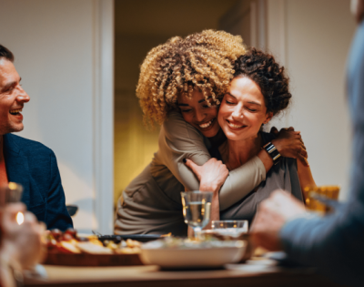 Two female friends at a dinner table looking happy, They are hugging. 