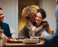 Two female friends at a dinner table looking happy, They are hugging. 